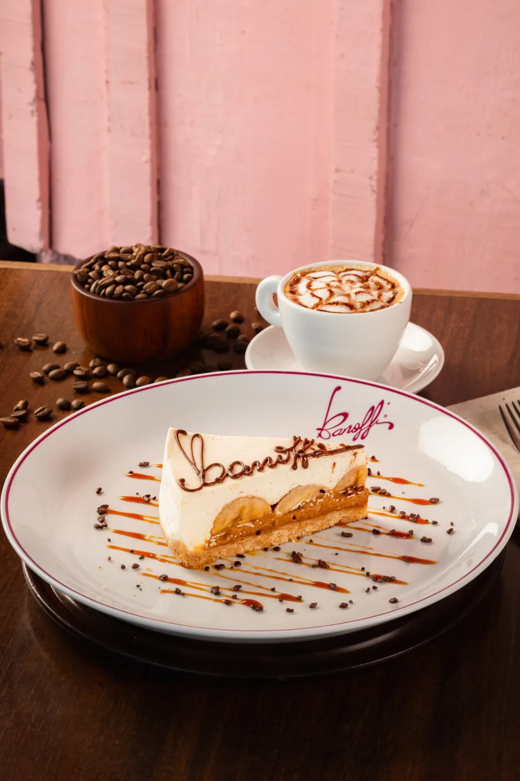 Fatia de Banoffi assinada em chocolate, no prato da casa, com cappuccino e a parede rosa ao fundo